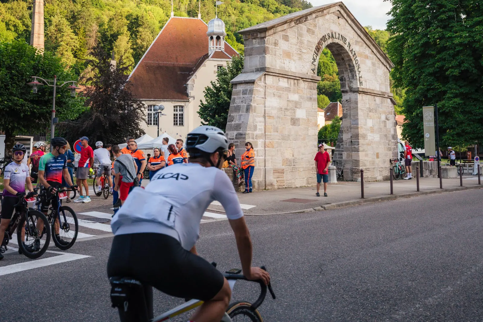 Un cycliste VELCAP à vélo de route devant l'arche de l'ancienne saline de Salins, d'autres cyclistes rassemblés au départ.